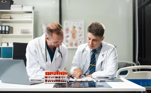 Two male doctors in white lab coats examining documents on a hospital desk, with medical charts and a laptop nearby.
