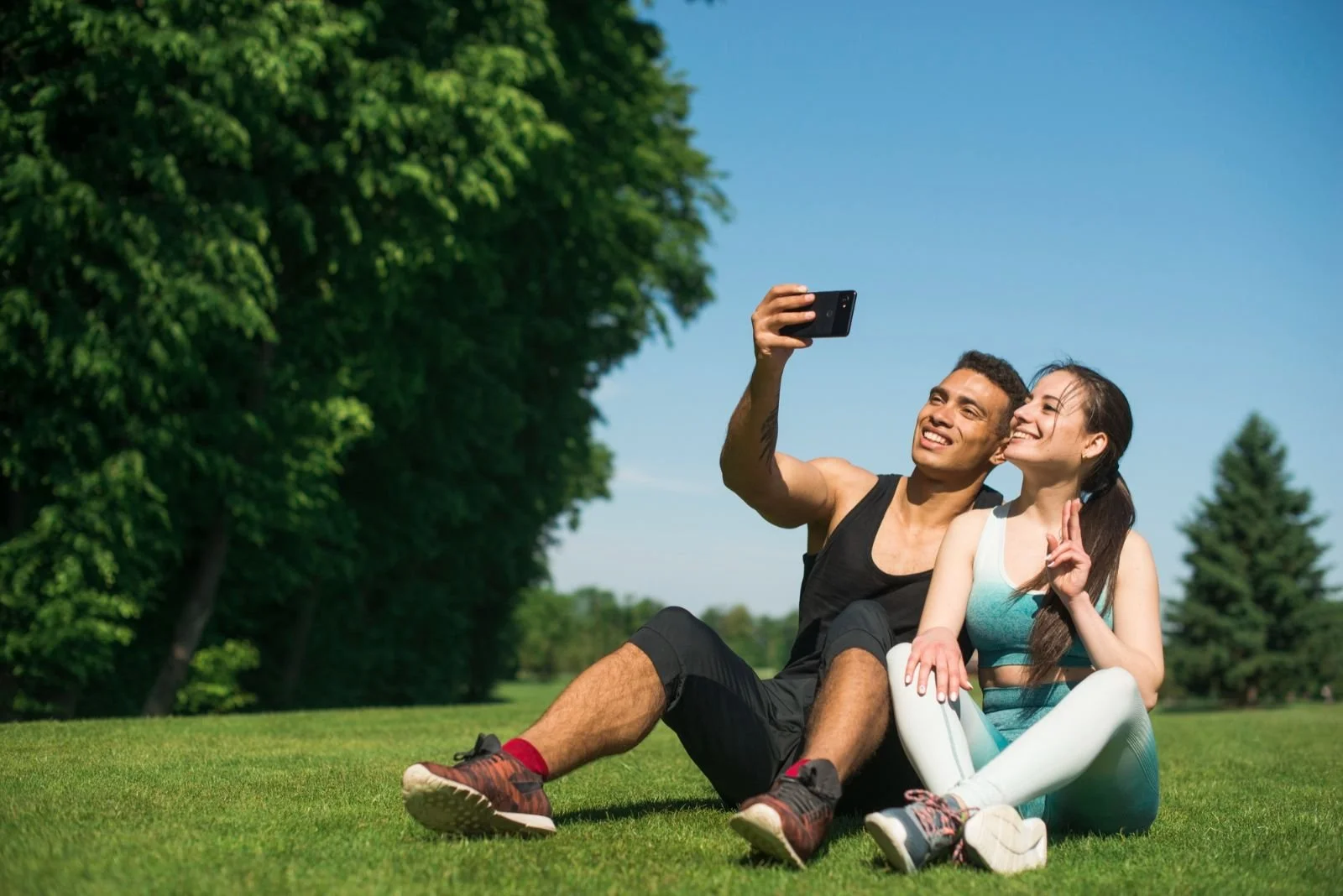 A young man and woman sitting on the grass in a park, taking a selfie together, smiling, with trees and a blue sky in the background.